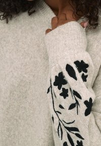 Pull en tricot beige clair avec un col côtelé et des broderies florales noires le long des manches. Texture douce, coupe ample.