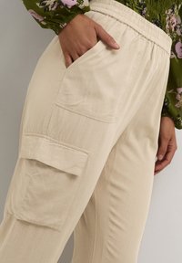 Béžové cargo kalhoty z lehkého materiálu, s elastickým pasem a boční kapsou s klopou. Hladký povrch, volný střih.