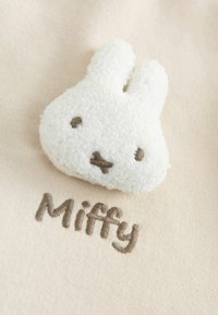 Biały, puszysty aplikowany kształt królika z brązowymi haftowanymi rysami twarzy na jasnobeżowym tle, z napisem "Miffy" wyhaftowanym poniżej.