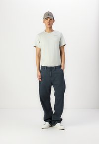 Grå caps, lys grønn t-skjorte, blå løstsittende jeans og hvite sneakers. Glatt stoff med en subtil logo-detalj på t-skjorten.
