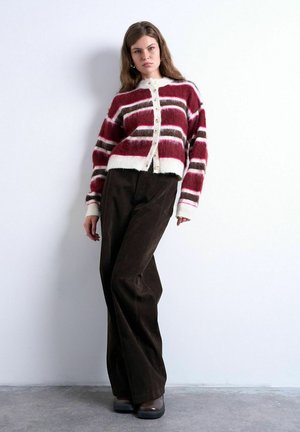 Femme portant un cardigan en maille duveteuse rayé bordeaux, blanc et marron, et un pantalon large marron foncé, debout contre un mur blanc.