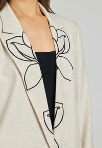 Blazer beige con ricami floreali neri indossato sopra una maglietta nera, che mostra il torso superiore e il collo su uno sfondo neutro.