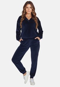 Marineblauer Velours-Trainingsanzug mit Top im V-Ausschnitt, langen Ärmeln und Hose mit elastischem Bund. Verfügt über seitliche Taschen und gerippte Bündchen. Klare Textur.