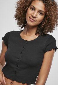 T-shirt noir court en tissu côtelé, avec un col rond, des manches courtes volantes et cinq boutons sur le devant.