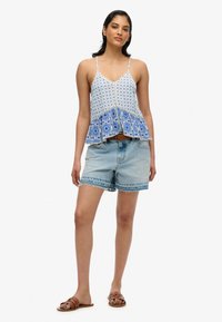 Top sin mangas blanco con acentos de patrón azul, dobladillo acampanado y tirantes ajustables, combinado con pantalones cortos de mezclilla azul claro. Sandalias marrones.