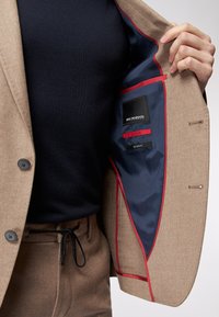 Brauner Blazer mit marineblauer Fütterung, roten Akzenten und Etiketten. Getragen über einem marineblauen Hemd. Hat Knöpfe und eine taillierte Passform.