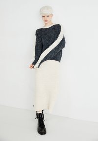 Maglione a maglia grigio scuro con striscia crema, abbinato a una gonna a maglia crema. Il modello indossa stivali neri con lacci e un cappellino chiaro.