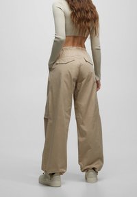 Beige wijde broekspijpen met een ontspannen pasvorm, twee achterzakken en een elastische tailleband. Materiaal lijkt glad met een subtiele glans.