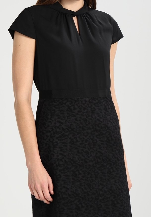 Femme portant une robe noire à manches courtes avec un décolleté en forme de goutte et une jupe à motifs texturés.