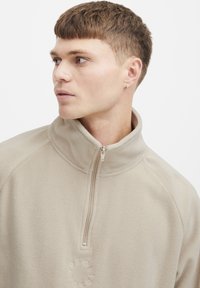 Light beige fleece pullover with a high collar, half zip, and logo detail on the chest. Soft texture, casual design with streamlined fit.