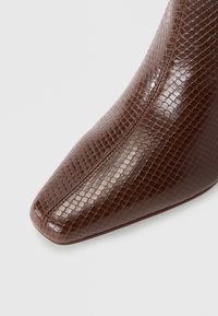 Gros plan d'une chaussure en cuir marron avec une texture brillante et écailleuse ainsi qu'une couture détaillée à l’avant.