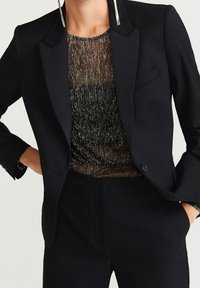 Blazer nero sartoriale con finitura in satin, abbinato a un top in maglia metallica trasparente. L'outfit presenta spalle strutturate e una vestibilità slim.
