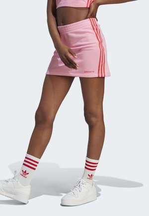 Femme portant une mini-jupe Adidas rose avec des rayures rouges, des chaussettes assorties et des baskets blanches, debout avec une main sur la hanche.