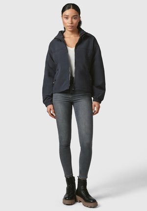 Marineblauwe lichtgewicht jas met een rits, elastische manchetten en twee voorzakken, gecombineerd met hoog getailleerde grijze jeans en zwarte veterschoenen.