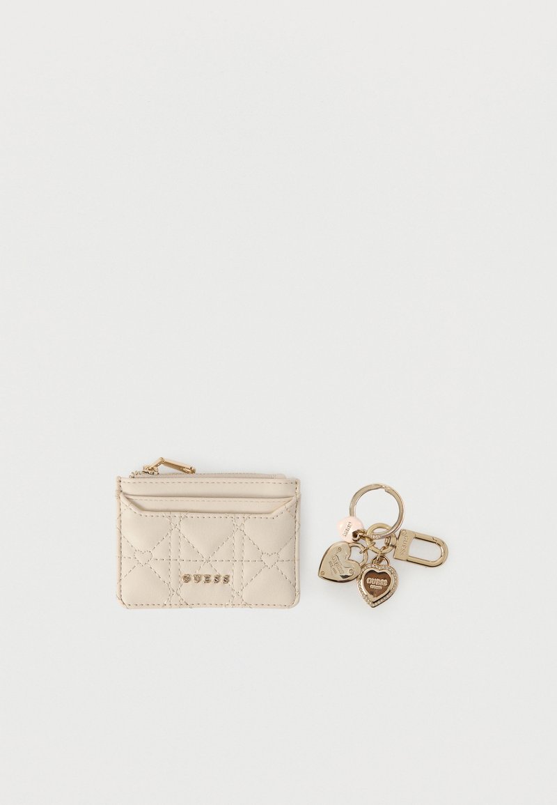 Porte-cartes matelassé beige avec logo Guess doré et porte-clés doré orné de breloques Guess en forme de cœur et de rond.