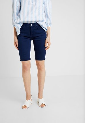 Persona con pantalones cortos azul marino hasta la rodilla, blusa a rayas azul claro y blanca, y sandalias blancas de tacón con la punta abierta, de pie frente a un fondo liso.