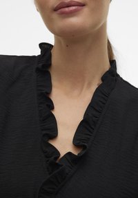 Blusa negra con un escote volante y tejido texturizado. El diseño incluye un corte en forma de V y un acabado liso alrededor del cuello.