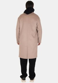 Cappotto di lana beige con taglio dritto, dettaglio della cucitura posteriore, abbinato a pantaloni in corduroy neri e una sciarpa nera texturizzata.