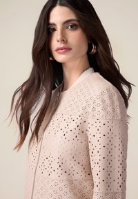 Veste perforée rose clair avec découpes florales, fermeture éclair à l'avant et col côtelé. Texture lisse avec une silhouette ajustée et des manches longues.