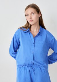 Niebieska koszula z długim rękawem, wykonana z lekkiego materiału, z kieszenią na piersi, luźnym krojem i elastycznym pasem.