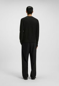 Mann, der sich abwendet, trägt einen strukturierten schwarzen Pullover, lockere schwarze Hosen und schwarze Schuhe vor einem schlichten hellgrauen Hintergrund.