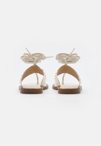 Sandali in pelle marrone chiaro con suola piatta, borchie dorate lungo i bordi e una cinghia annodata color crema alla caviglia, che mettono in mostra un design aperto sul retro.