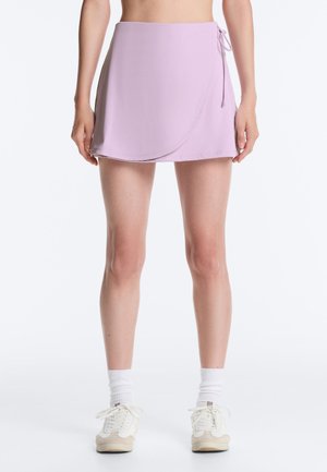 Žena oblečená v fialovej zavinovacej mini sukni so stranovým viazaním, biele členkové ponožky a biele tenisky, stojí pred jednoduchým pozadím.