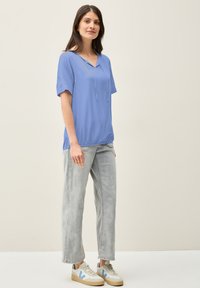Top azzurro chiaro a maniche corte con scollatura a V e dettaglio con cordino, abbinato a jeans a gamba larga grigio chiaro e scarpe da ginnastica bianche con accenti blu.