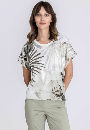 Vrouw met schouderlang golvend haar, gekleed in een wit shirt met korte mouwen en grijs-beige blad- en bloemenprints, en lichtgroene broek.
