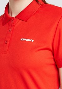 Punainen polo-paita, joka on valmistettu sileästä kankaasta ja jossa on ribattua kaulusta. Siinä on kolme nappia ja valkoinen "ICEPEAK" -logo vasemmassa rintataskussa.