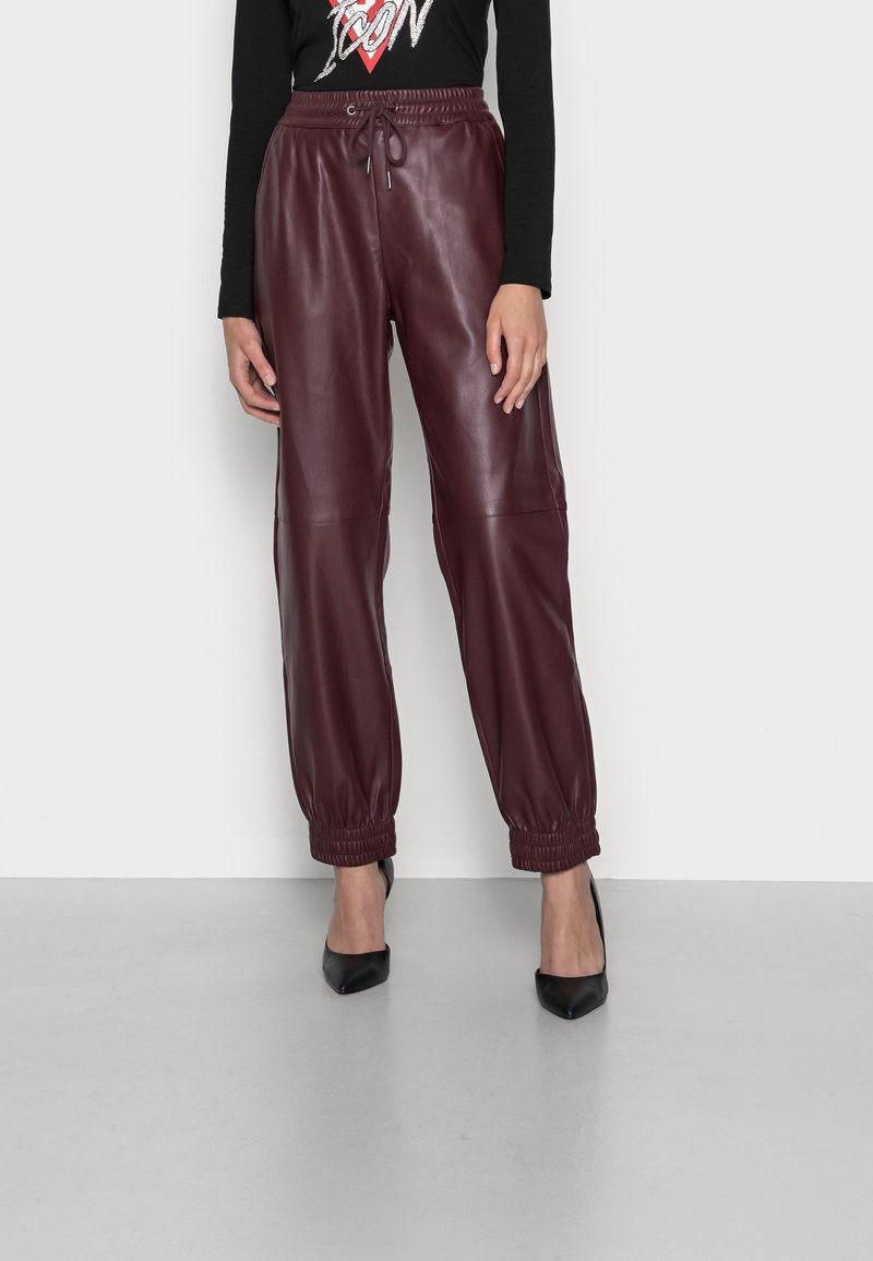 Femme portant un pantalon jogger en cuir bordeaux taille haute avec des poignets élastiques, associé à des talons noirs pointus et un haut noir à manches longues.
