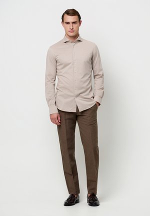 Camicia beige chiaro con bottoni e maniche lunghe, abbinata a pantaloni marroni. Vestibilità slim con una texture liscia e una lucentezza sottile. Scarpe nere completano il look.