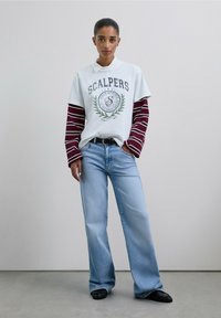 Mujer de pie con la mano en el bolsillo, vistiendo una camiseta blanca de "Scalpers" sobre una camiseta de mangas largas a rayas color burdeos, jeans anchos de color azul claro y zapatos negros.