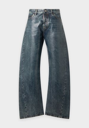 Femme portant une veste en cuir marron, un t-shirt noir, un jean large et foncé, des chaussures noires pointues et une casquette de baseball noire sur un fond uni.