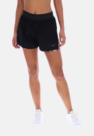 Kvinde iført sorte træningsshorts med farverig indvendig foring og hvide sneakers, stående mod en ensfarvet baggrund.