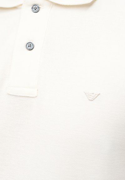Λευκό πόλο πουκάμισο με δύο μαύρα κουμπιά και μικρό κεντημένο λογότυπο στην αριστερή πλευρά του στήθους.