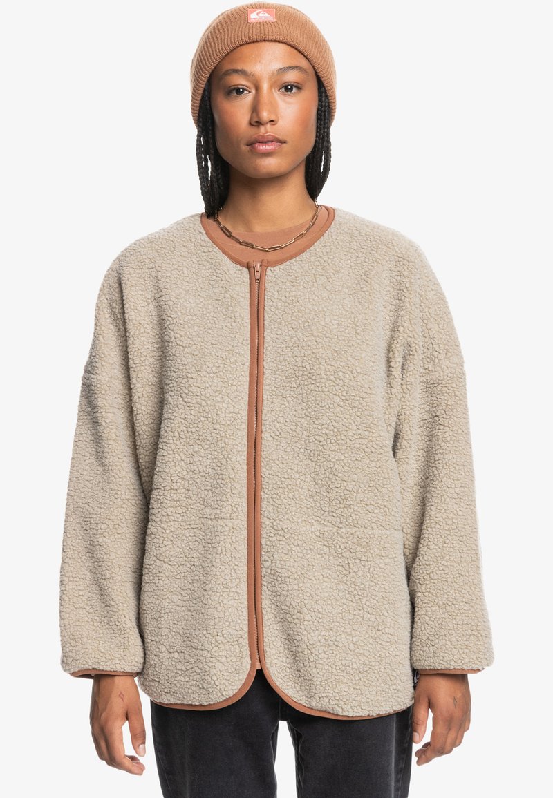 Beige fleecejacka med rundade kanter, kontrasterande brun dragkedja och trim. Långa ärmar, texturerad tyg och inga synliga mönster.