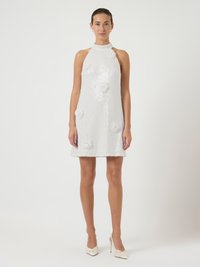 Γυναίκα που φοράει ένα λευκό αμάνικο φόρεμα με ασημώματα και λουλουδάτες εφαρμογές, συνδυασμένο με λευκά ψηλά τακούνια, που στέκεται μπροστά από έναν απλό φόντο.