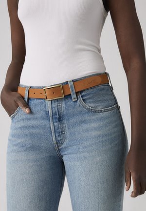 Persona dalla pelle scura che indossa jeans azzurri chiari con una cintura di pelle marrone con fibbia dorata e una canottiera bianca a coste senza maniche.