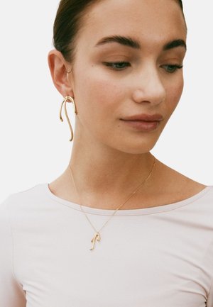 Gold-tone metal jewelry featuring a curved earring and a delicate pendant necklace with an abstract design, worn over a light pink top.