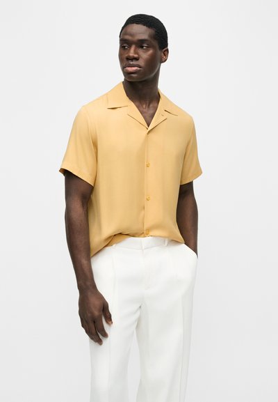 Μαύρος άνδρας που φοράει κίτρινο πουκάμισο με κοντά μανίκια και κουμπιά, μπασεμένο σε άσπρο παντελόνι με ψηλή μέση, στέκεται με το ένα χέρι στην τσέπη.