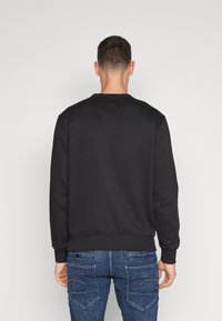 Muž ve jednoduché černé mikině a modrých džínách stojí zády k bílé stěně.