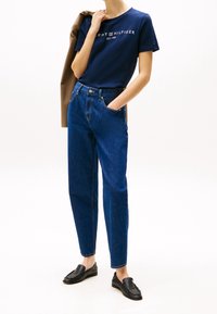 Modré džíny s vysokým pasem a volným střihem, doplněné tmavě modrým bavlněným tričkem s bílým logem. Černé mokasíny dokončují vzhled.