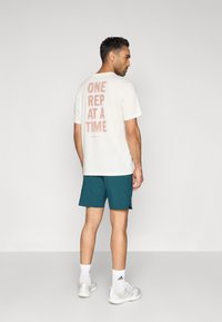 Λευκό βαμβακερό μπλουζάκι με τη φράση "ONE REP AT A TIME" τυπωμένη στην πλάτη σε ξεθωριασμένο κόκκινο διαβαθμισμένο χρώμα, συνδυασμένο με τιρκουάζ σορτς και λευκά αθλητικά παπούτσια.