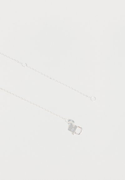 Collier délicat en chaîne d'argent avec un fermoir à ressort et une petite étiquette ovale près du fermoir sur fond blanc uni.