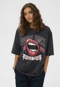 Göndör hajú nő sötét, túlméretezett pólót visel, amelyen egy piros, nyitott száj grafika és a "Rebel Rock Tour 1990 World Tour" felirat látható.