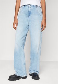 Person som bär ljusblå vida jeans och svarta kängor med tjock sula, stående mot en enfärgad ljusgrå bakgrund.