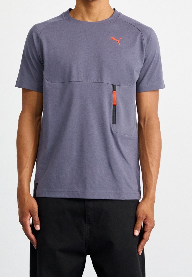 Homme portant un t-shirt gris à manches courtes Puma avec un logo orange et une poche verticale zippée sur la poitrine.