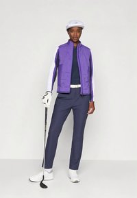 Lila gepolsterte Weste über einem marineblauen Hemd, kombiniert mit marineblauen Hosen und weißen Schuhen. Das Outfit umfasst eine weiße Golfmütze und einen Golfschläger.