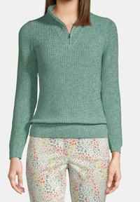 Donna che indossa un maglione a zip in maglia turchese e pantaloni bianchi con un piccolo motivo floreale multicolore, in piedi davanti a uno sfondo semplice.